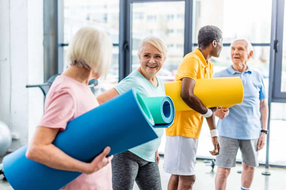 senior sportswomen holding fitness mats and their male friends standing behind at gym, independent living