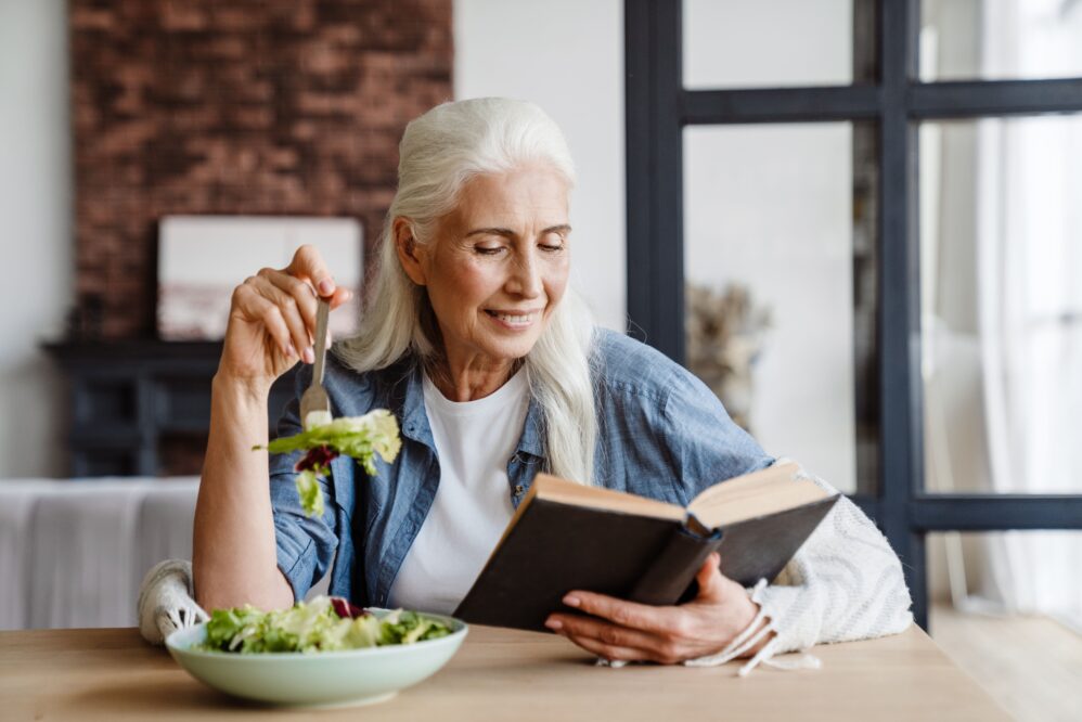 Smiling senior woman reading a book while sitting in the kitchen, eating salad