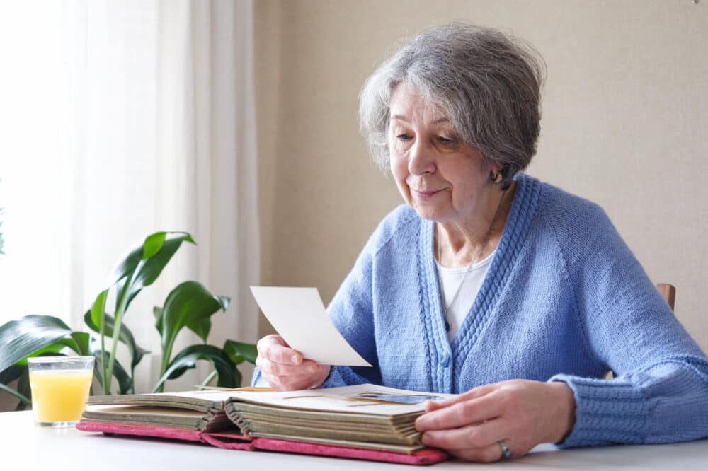 An elderly woman with nostalgia looks at old photos in a photo album or photobook - The concept of family and life values and loneliness in old age