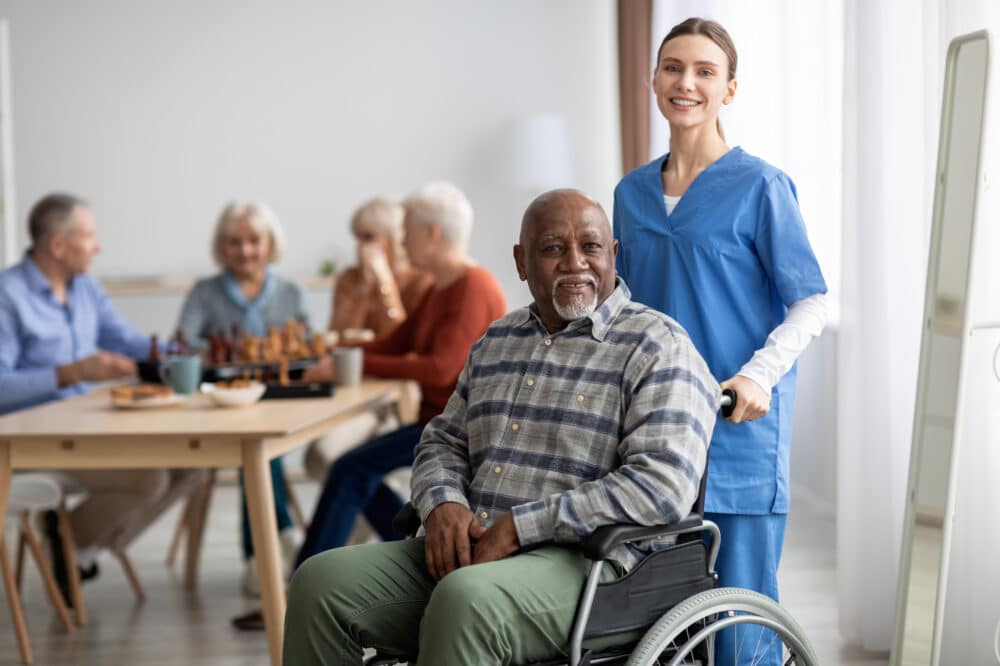 Happy black man older patient on wheelchair with female nurse smiling at camera, group of senior people sitting on couch on background