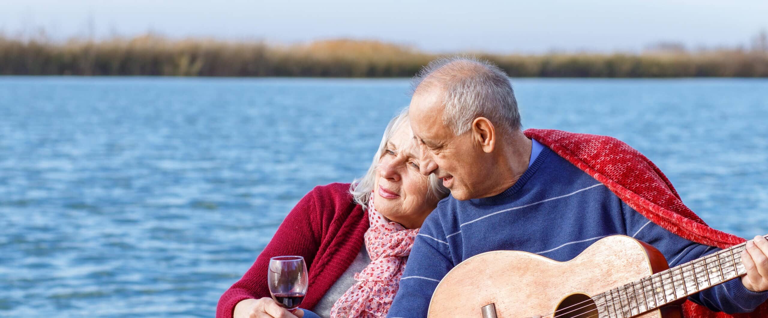 Happy senior couple enjoying time together playing guitar and drinking wine by the lake wrap around in a red blanket.