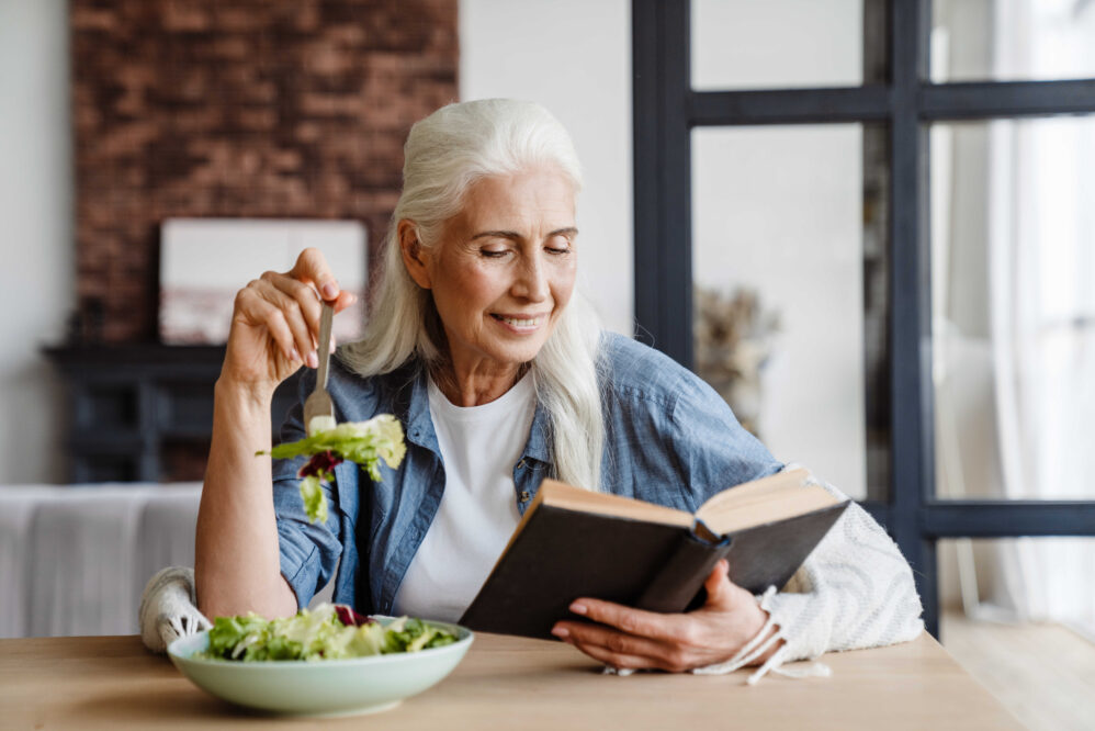 Smiling senior woman reading a book while sitting in the kitchen, eating salad