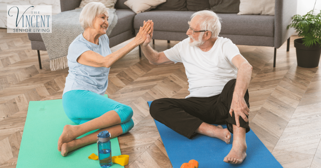 Active senior couple smiling and giving a high five during a seated exercise session on yoga mats, promoting memory care for elderly through physical activity and social engagement.