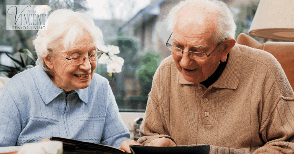 Two memory care residents smiling while looking through a photo album together, seated in a cozy, sunlit room