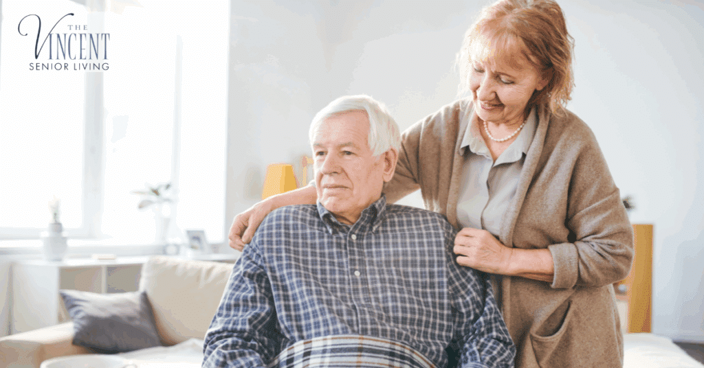 A woman gently comforting her husband, a memory care resident, as he sits wrapped in a blanket in a bright, comfortable room