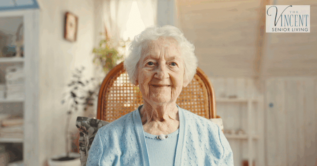 An elderly woman with white hair, wearing a light blue cardigan, smiles warmly while sitting in a bright, cozy room, representing compassionate dementia care for the elderly.