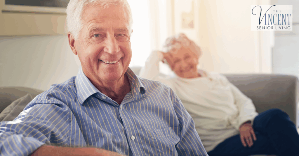 An elderly man in a striped shirt smiles from a sofa with an elderly woman relaxed in the background, illustrating the comfort and support offered through dementia care for the elderly.