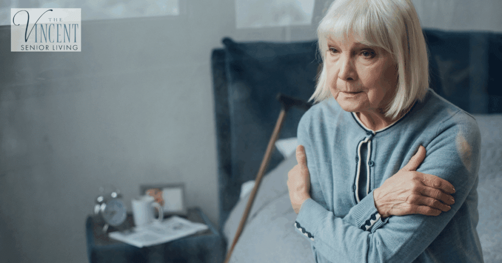 Alzheimer’s care at home: elderly woman sitting on her bed with arms crossed, looking thoughtful, cane nearby in a quiet bedroom.