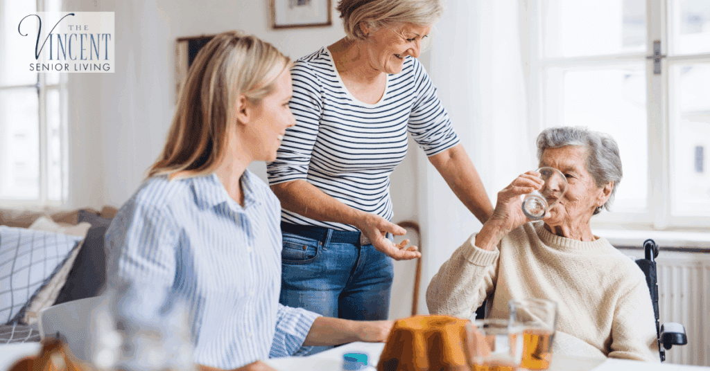Alzheimer’s care at home: senior woman in a wheelchair drinking water while two family care partners support her at a bright kitchen table.