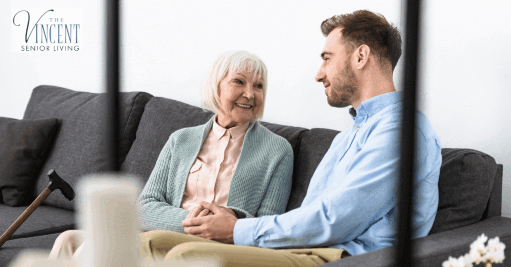 Visiting a loved one in memory care — a smiling elderly woman sitting on a couch, holding hands and talking warmly with a younger man.