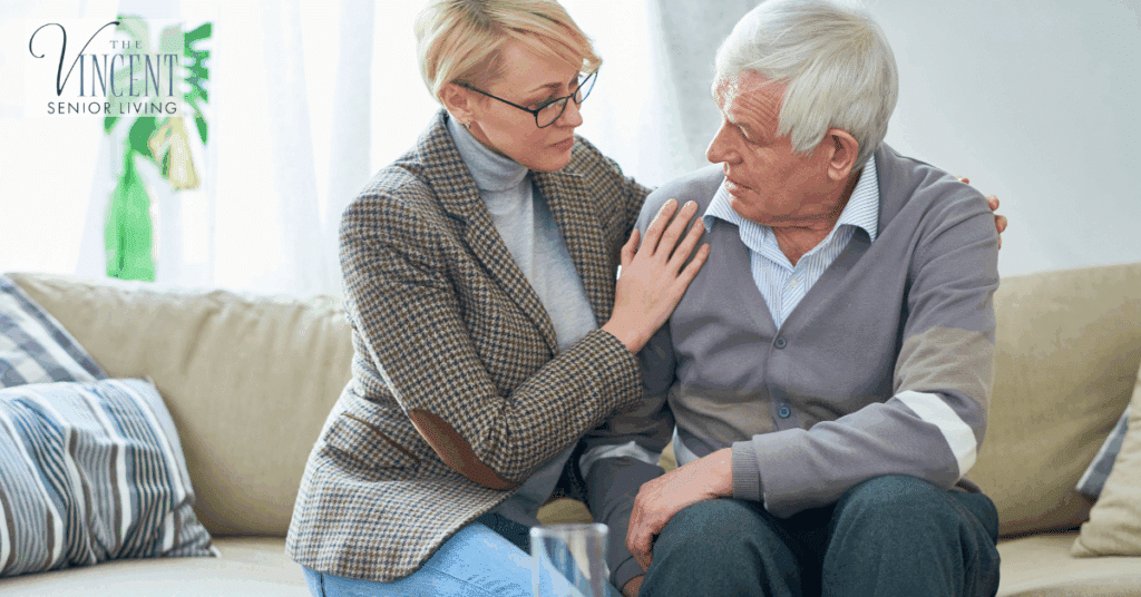 An older man sitting on a couch receiving comforting support from a woman as they talk, illustrating a compassionate senior memory care setting.